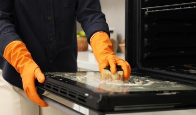 pessoa com luvas de limpeza laranja esfregando um fogão com uma esponja - limpeza da cozinha - Assaí Atacadista