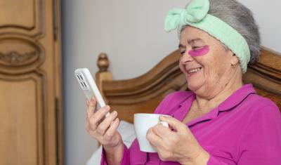 senhora branca segurando um celular e uma xícara nas mãos sentada na cama com uma mascara nas olheiras e uma faixa no cabelo - produtos de autocuidado - Assaí Atacadista