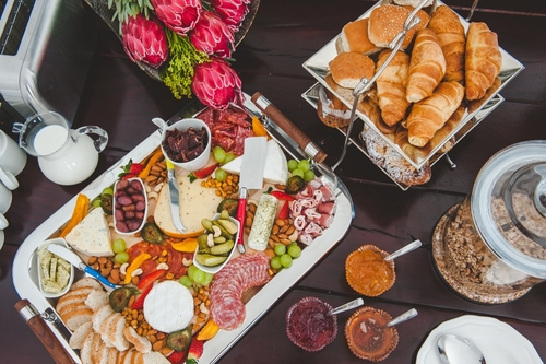 tábua de frios com diversos ingredientes acompanhada de potes de geleia, um cesto de pães e um pequeno buquê de flores - Assaí Atacadista